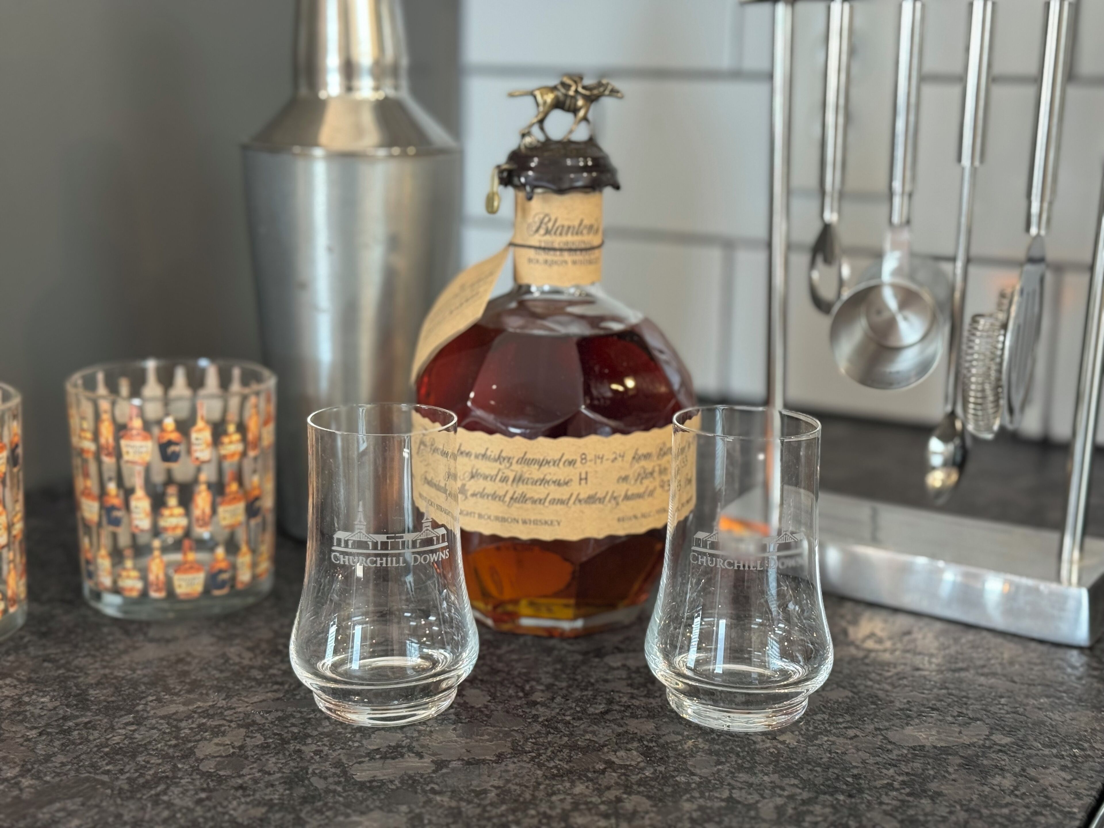 Uma garrafa de uísque bourbon Churchill Downs, dois copos medidores e um castiçal decorativo em uma bancada de cozinha.