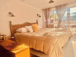 A bedroom with a wooden headboard, a bed with beige bedding, two wall-mounted lamps, and a view through a window with curtains.