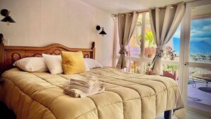 Una cama con cabecera de madera, vista de un paisaje urbano a través de una ventana y un balcón con mesa.