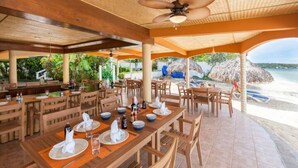 Un restaurant situé en bord de mer, avec des tables et des chaises en bois, un ventilateur de plafond et une vue sur la plage et l’océan.