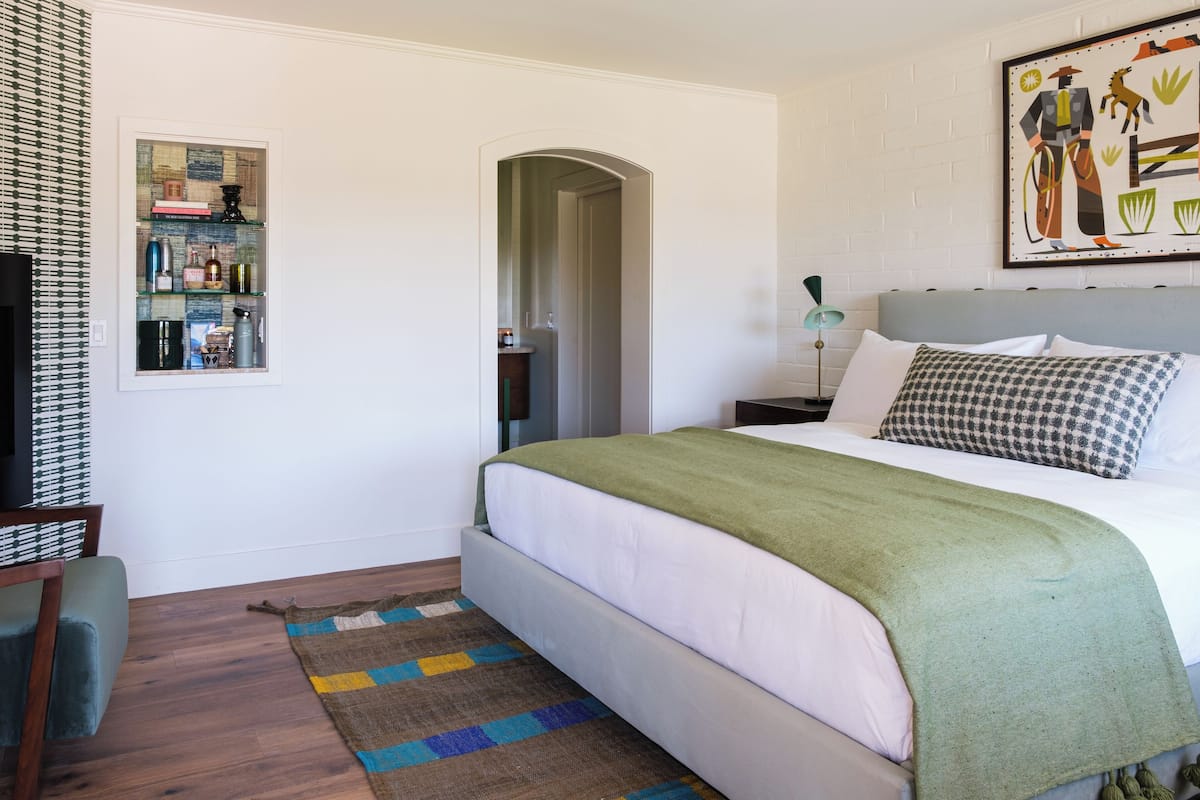A neatly arranged bedroom with a bed, a bedside table, a lamp, a chair, a rug, and a wall-mounted shelf with various items.