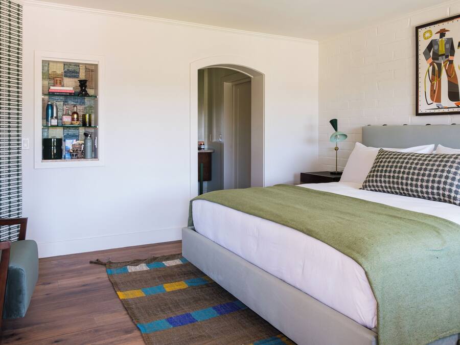 A neatly arranged bedroom with a bed, a bedside table, a lamp, a chair, a rug, and a wall-mounted shelf with various items.