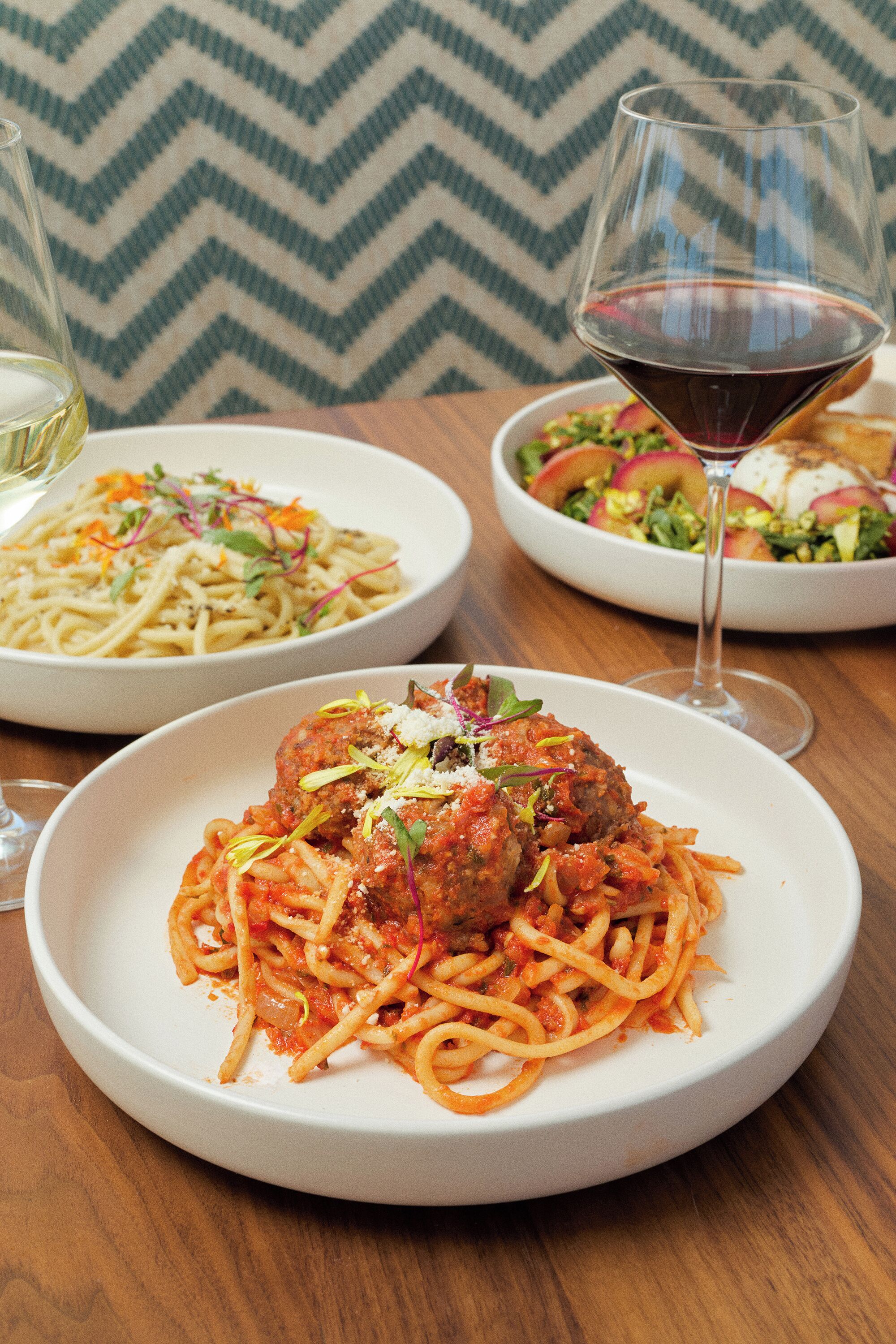 A table with three dishes: spaghetti with meatballs, a pasta salad, and a vegetable salad.