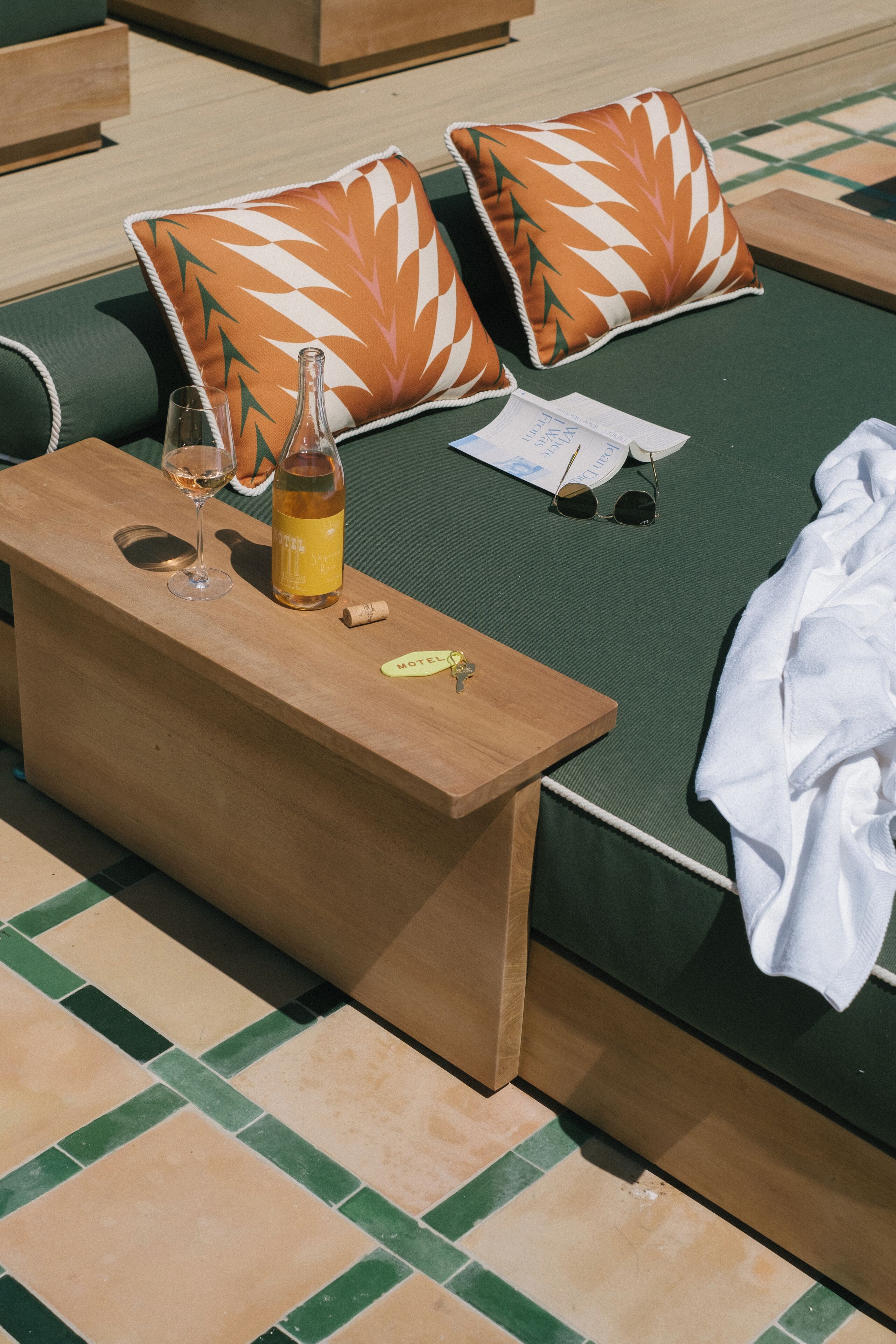 Ein sonnendurchflutetes Zimmer mit einem grünen Sofa, zwei orangefarbenen Kissen mit Blattmuster, einem Holztischchen mit einer Weinflasche, Gläsern und einem Handtuch.