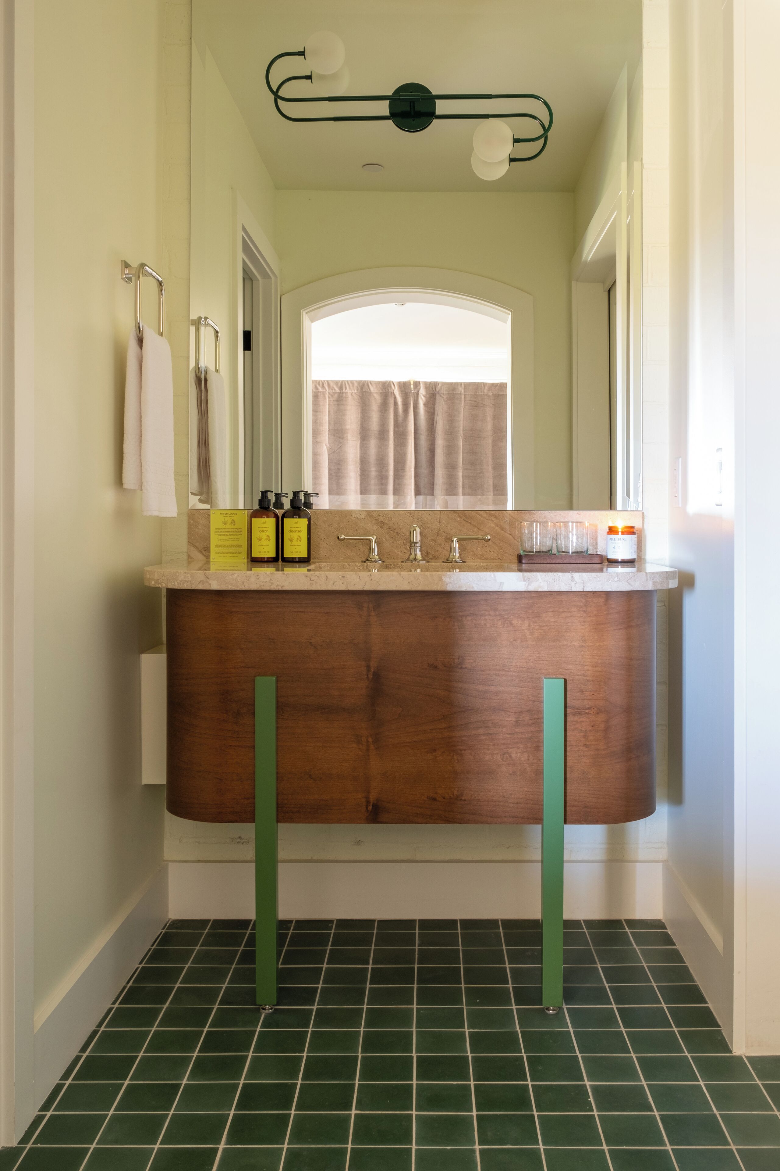 A bathroom with a wooden vanity, green floor tiles, a mirror, and a ceiling light fixture.