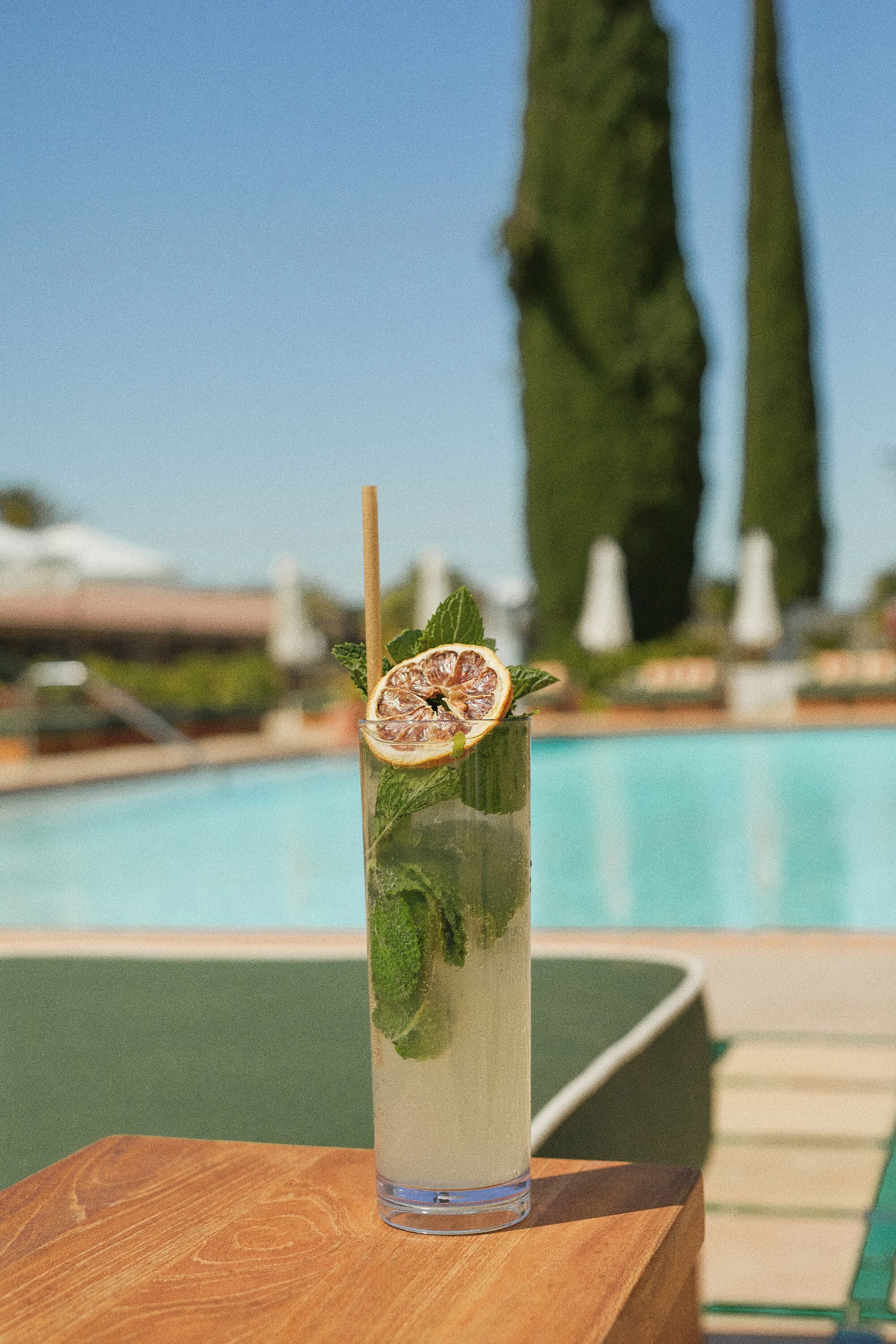 Un grand verre avec une paille, décoré d’une tranche d’agrumes et de feuilles de menthe, posé sur une surface en bois près d’une piscine.