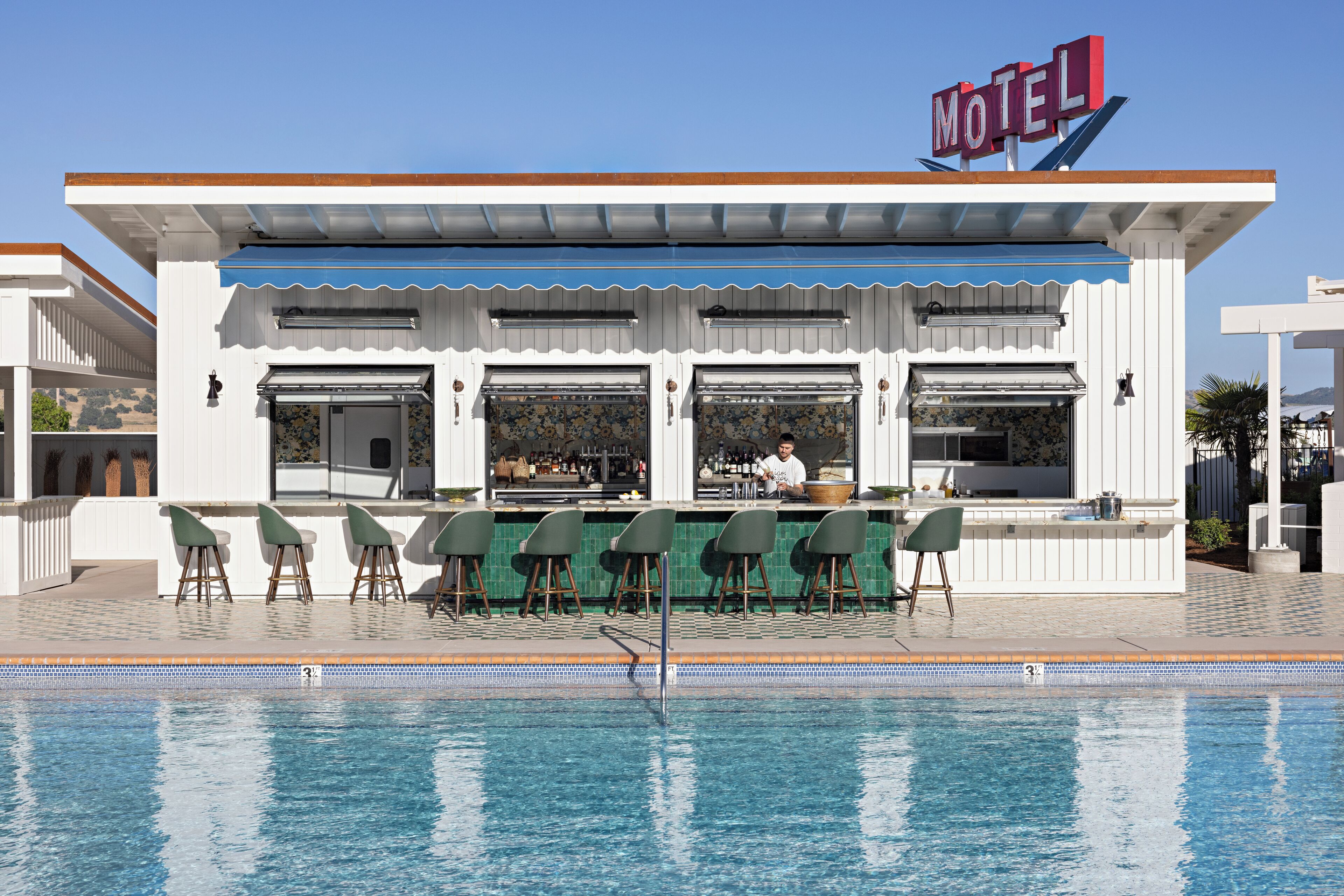Un bar junto a la piscina con un cartel de 'Motel', taburetes y un camarero.