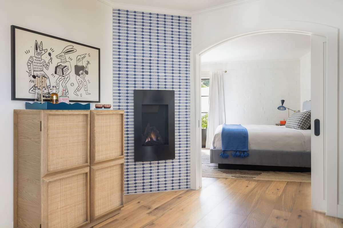 A modern bedroom with a fireplace, a bed with a blue blanket, a wooden wardrobe, and a patterned wall.