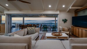 A modern living room with a large TV, comfortable seating, and a view of the ocean.