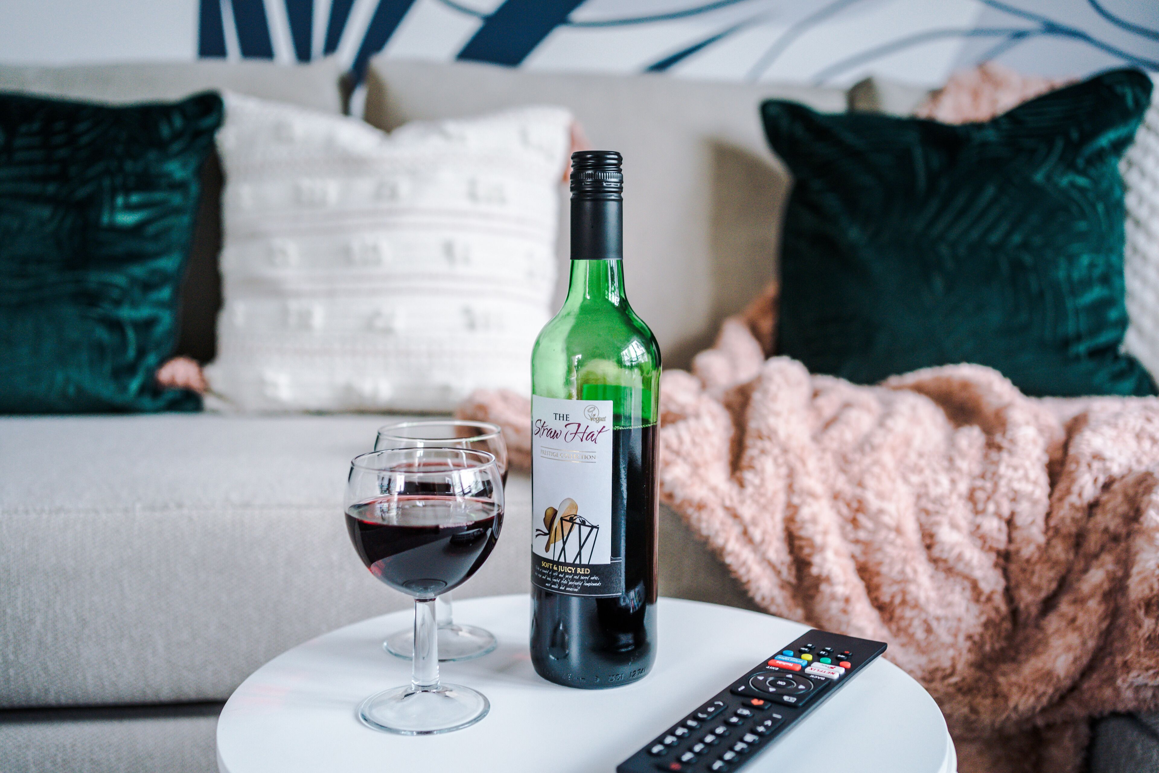A bottle of wine and a glass on a white table in a living room with a couch and pillows in the background.