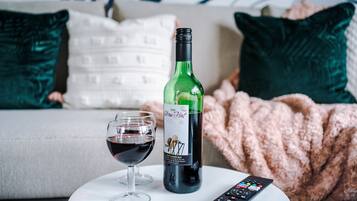 A bottle of wine and a glass on a white table in a living room with a couch and pillows in the background.