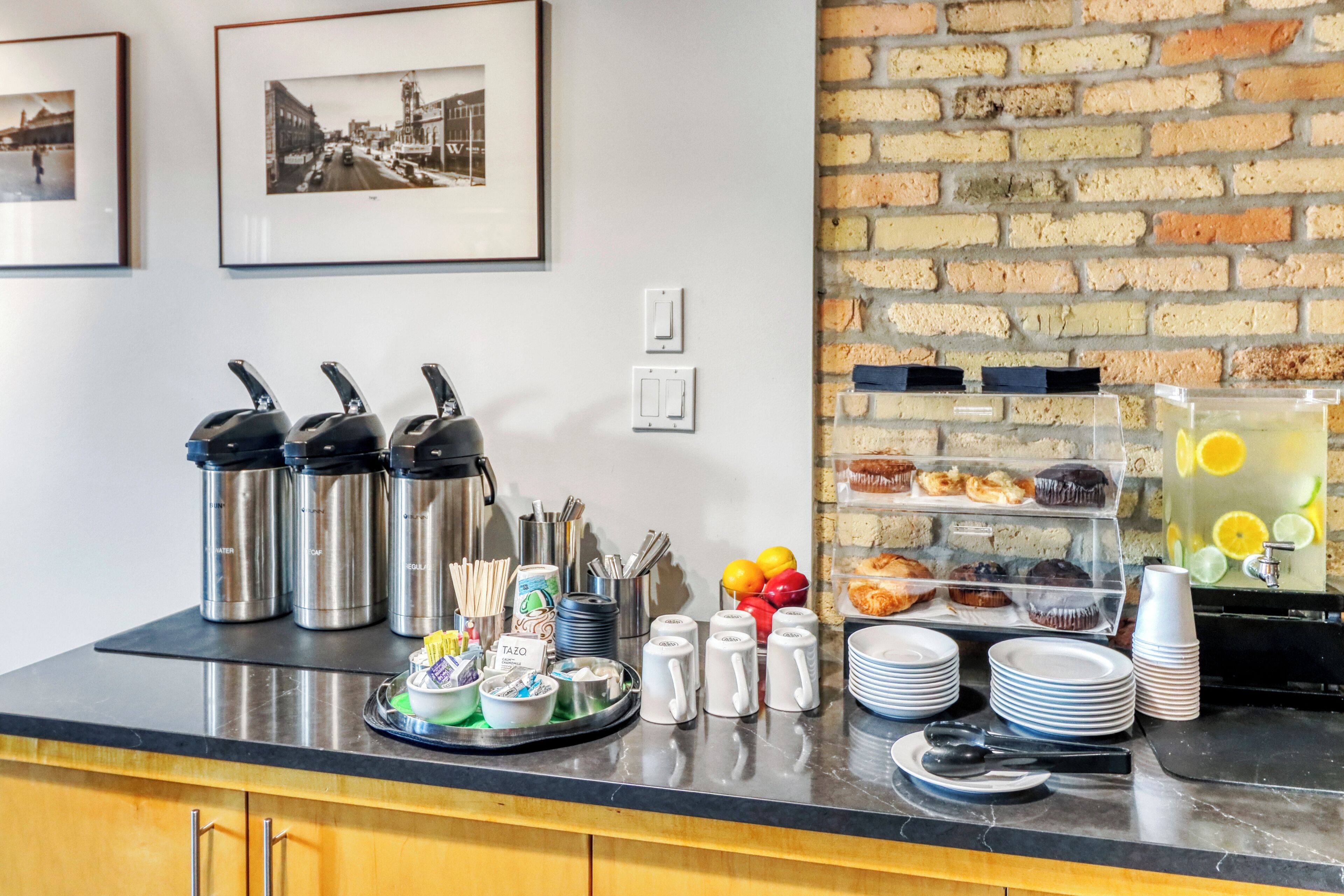 A breakfast buffet with coffee makers, mugs, plates, and a fruit display.