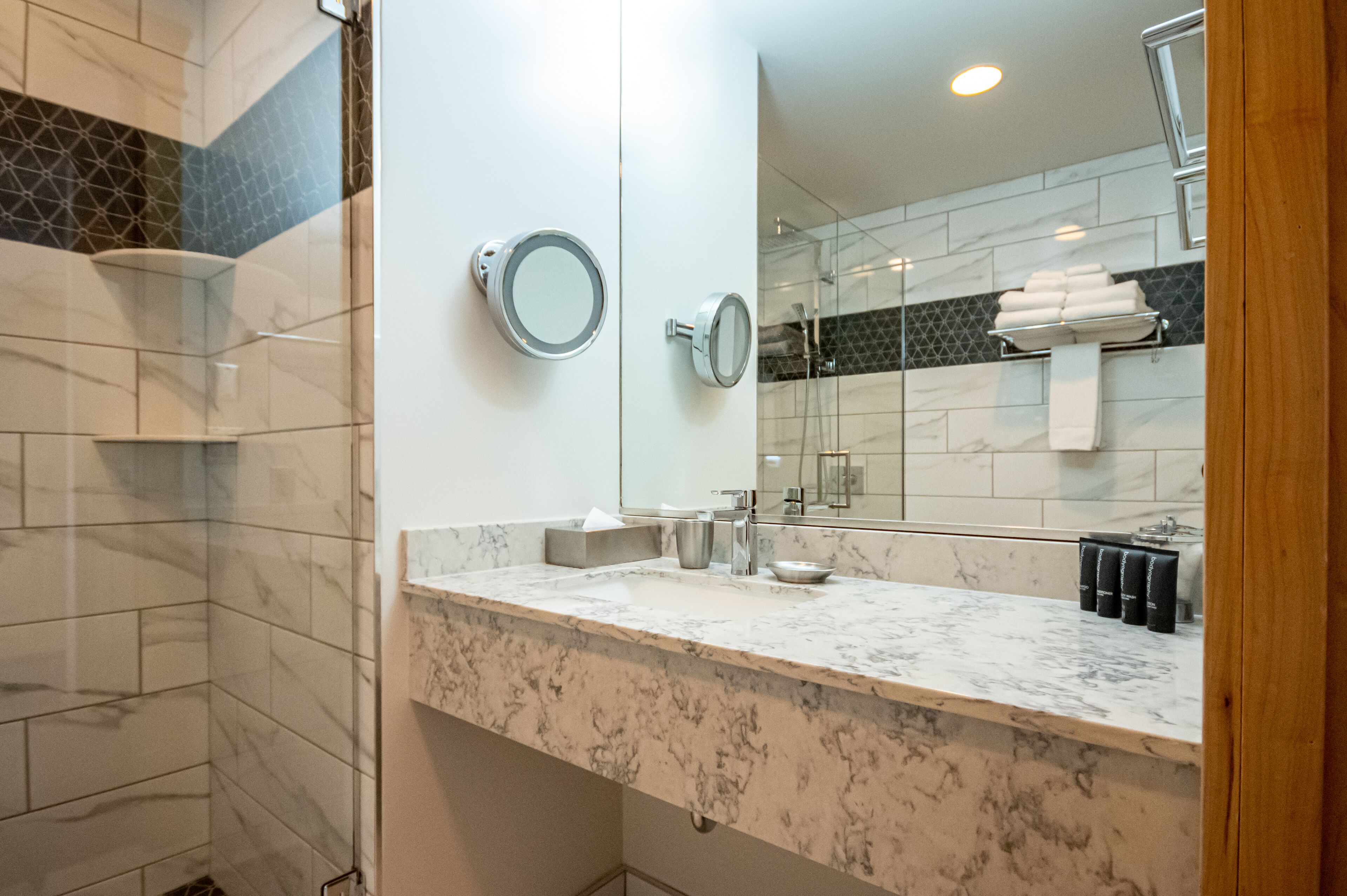A bathroom with a marble countertop, two round mirrors, a sink, and a glass shower enclosure.
