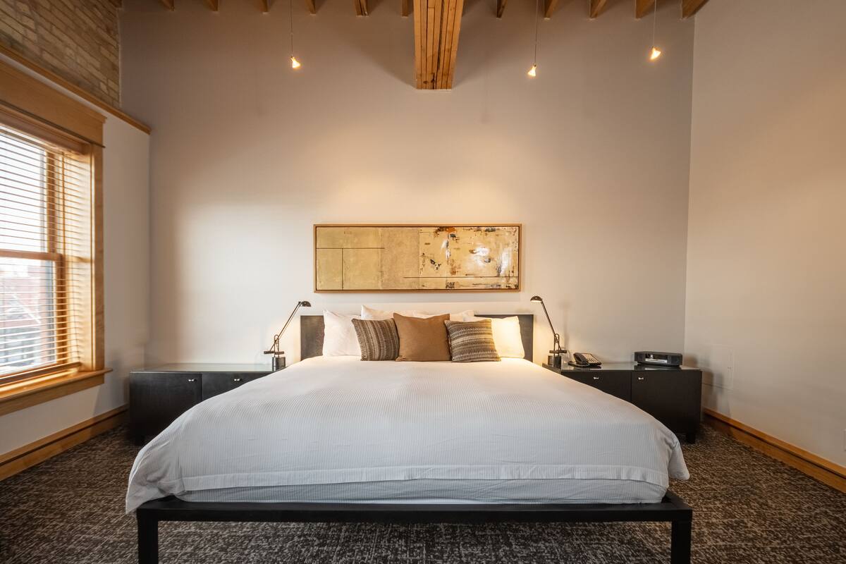 Une chambre avec un grand lit, des poutres apparentes en bois au plafond, un grand tableau abstrait et une fenêtre avec des stores.