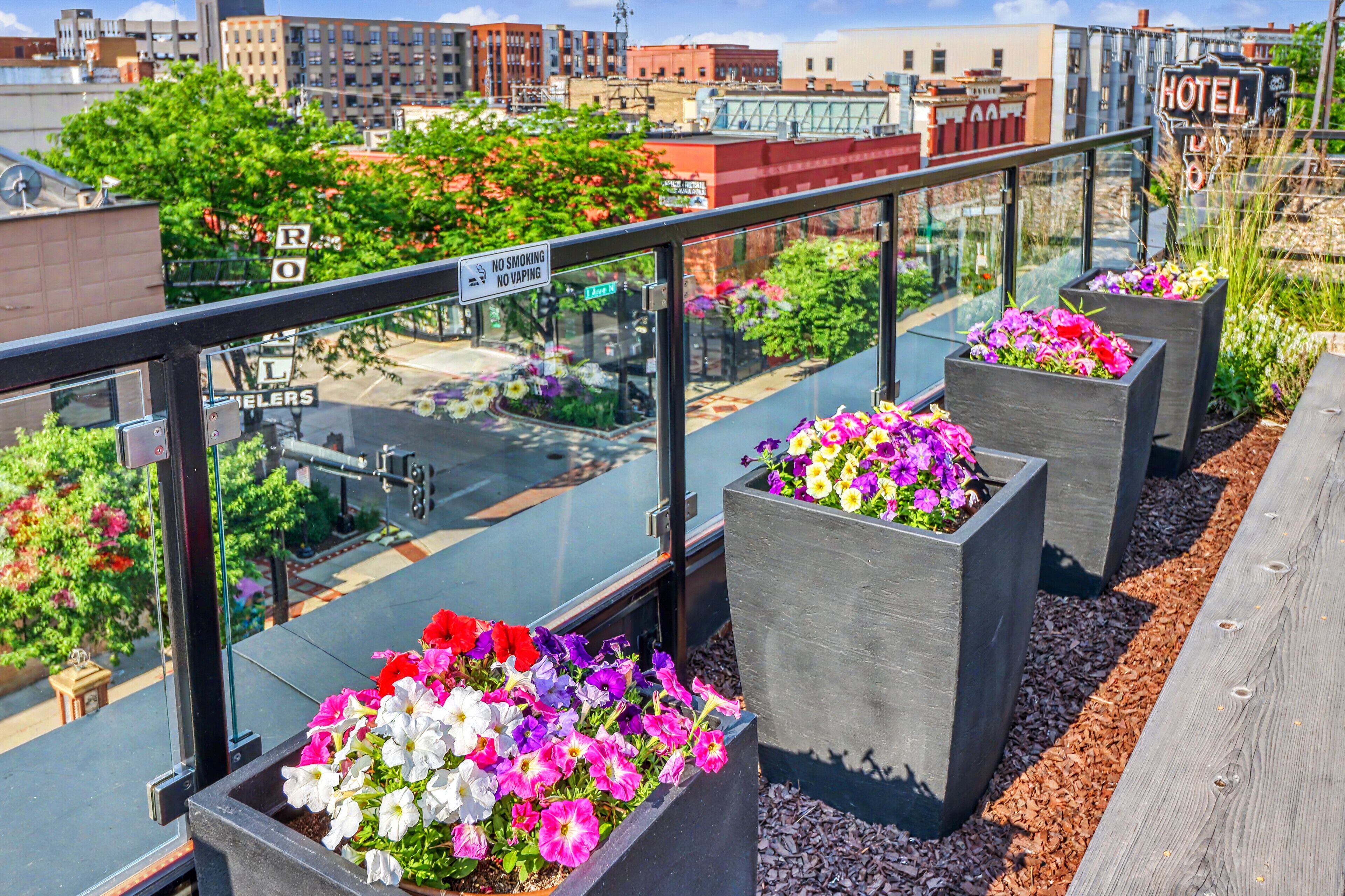 A rooftop terrace with flower planters and a glass railing overlooking a cityscape.