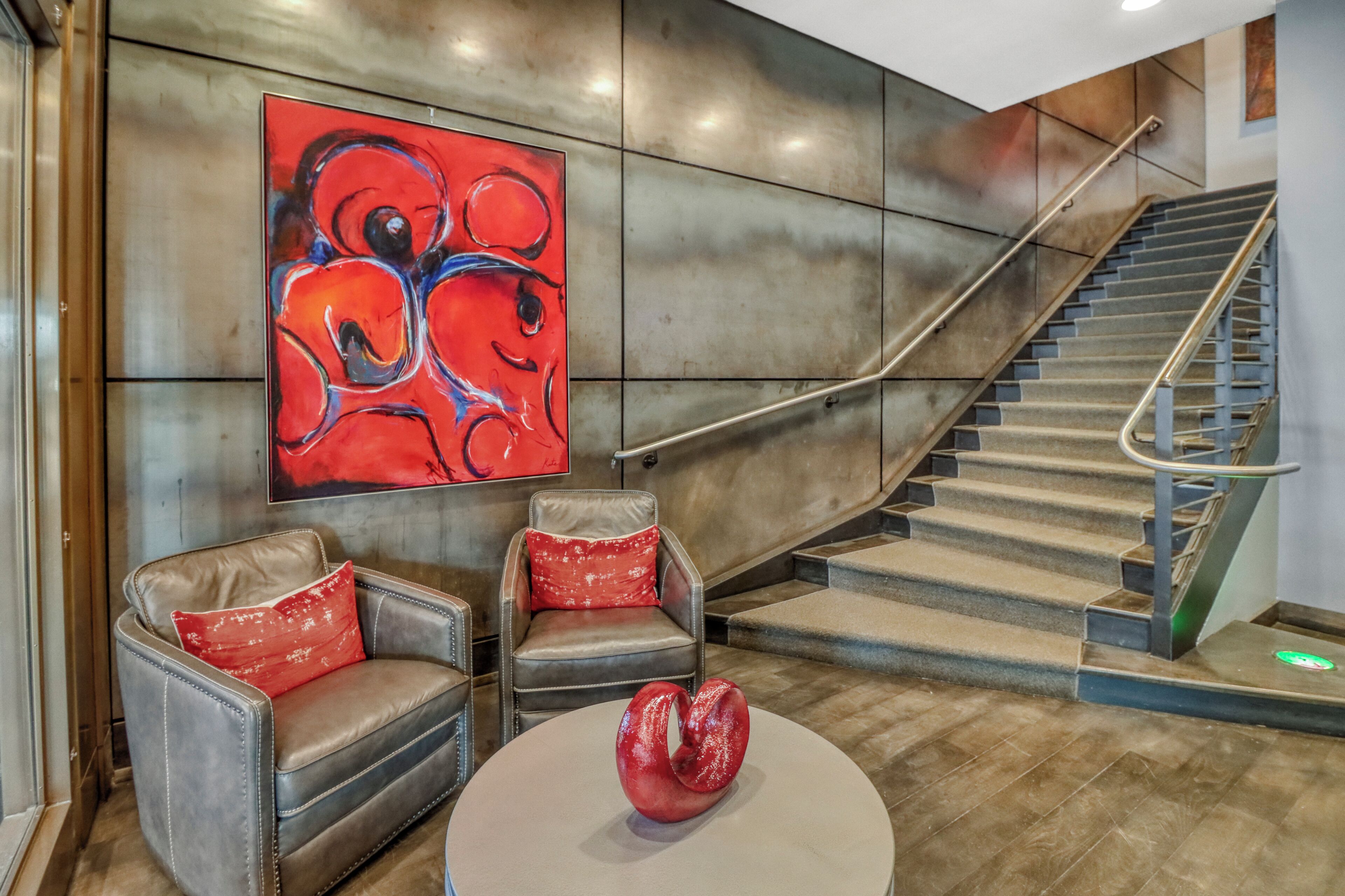 A modern lobby with a staircase, leather armchairs, a red abstract painting, and a unique red sculpture.