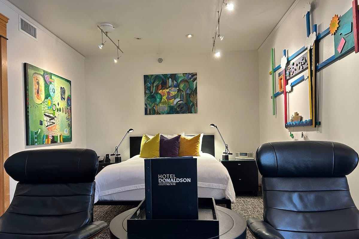 Une chambre d’hôtel avec un lit, deux fauteuils en cuir noir, une table ronde et des œuvres d’art accrochées aux murs.