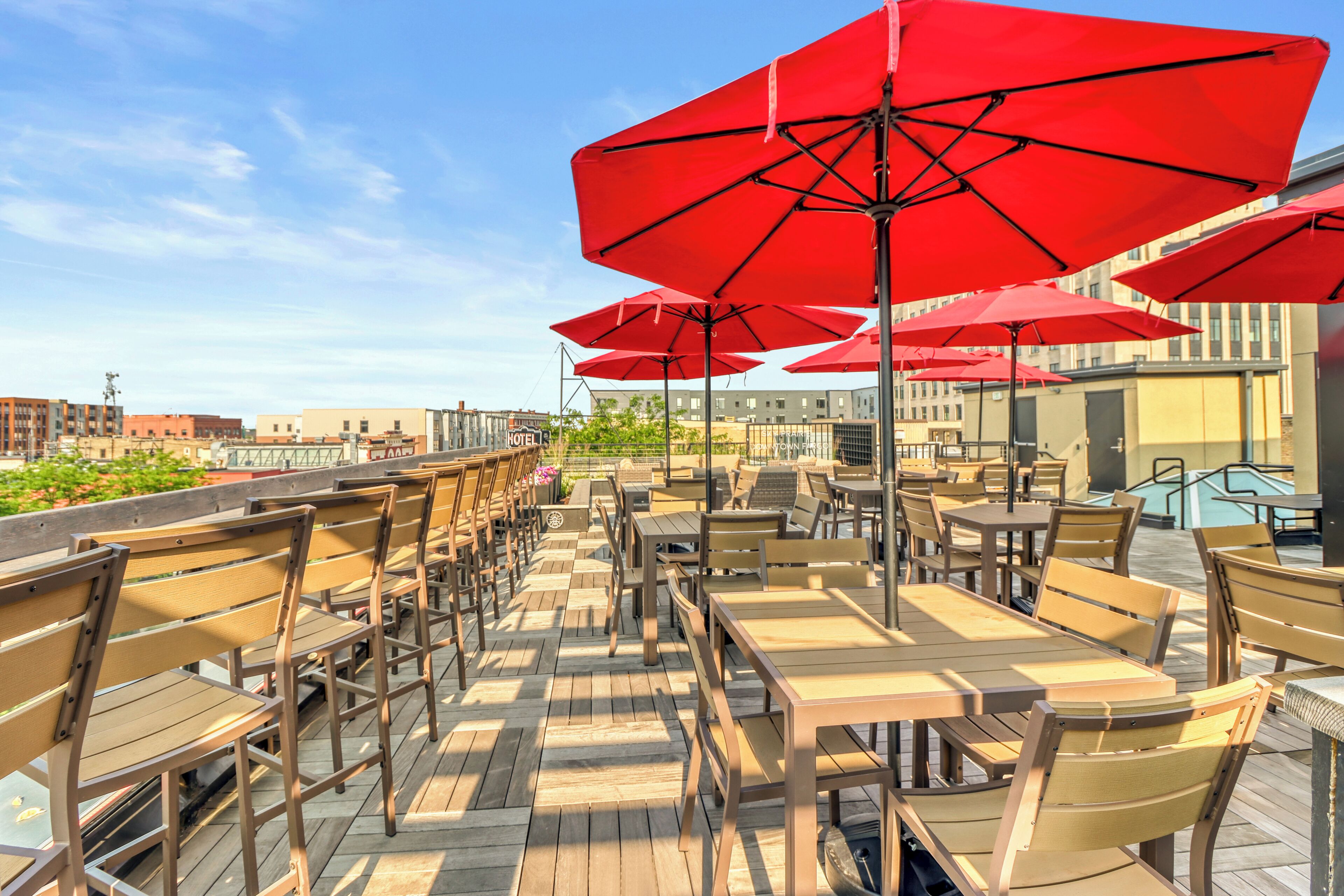 An outdoor seating area with wooden tables and chairs, red umbrellas, and a view of buildings in the background.