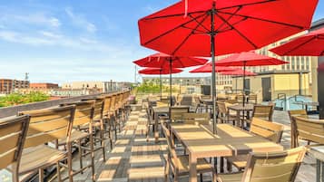 An outdoor seating area with wooden tables and chairs, red umbrellas, and a view of buildings in the background.