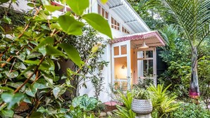A white house with a red roof, surrounded by lush greenery and a garden with various plants and a bird bath. - Flower of Life (Limon Province)