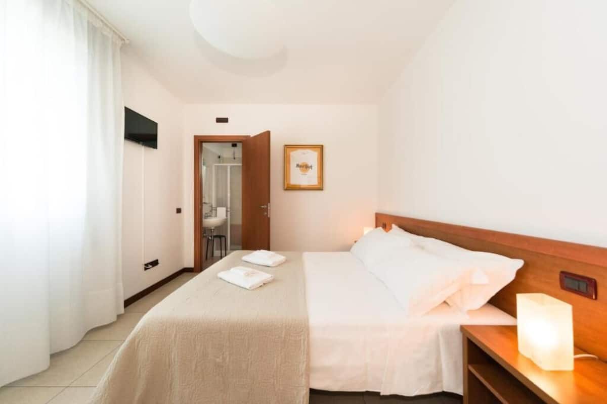 Une chambre d’hôtel avec un lit, une table de chevet, une lampe de chevet, un tableau encadré au mur et une salle de bain visible à travers une porte ouverte.