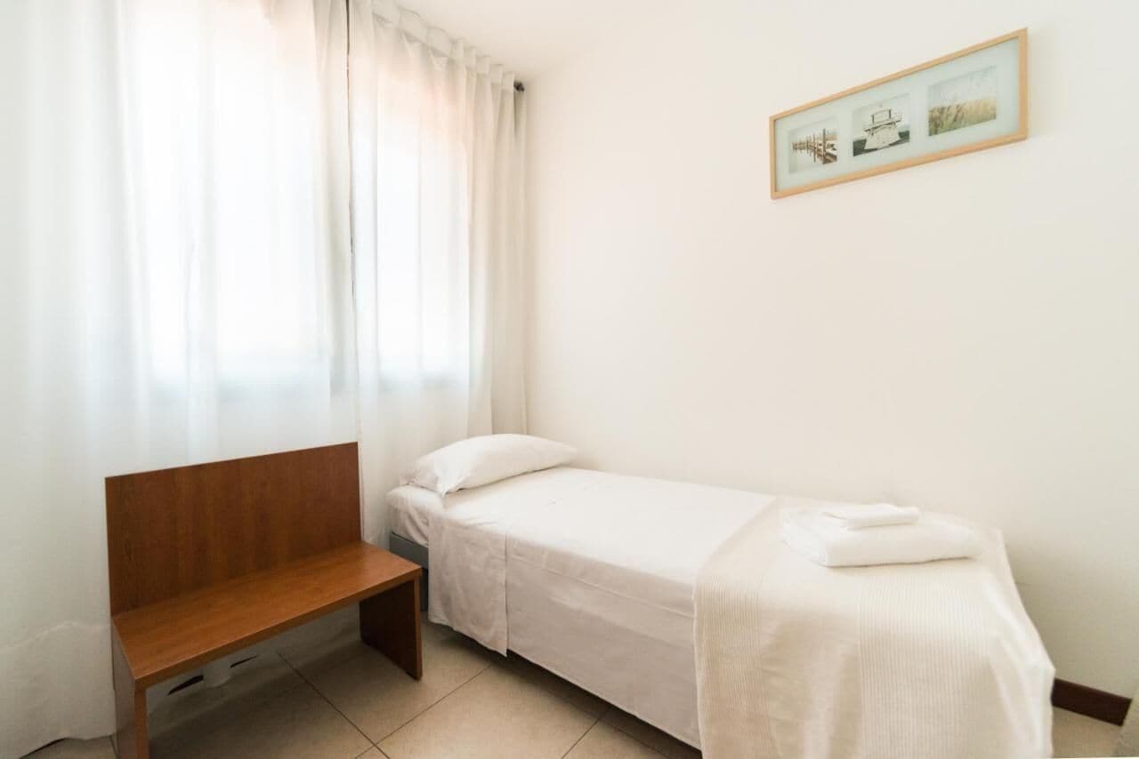A small, single-bed room with a wooden bench, a window with sheer curtains, and a framed picture on the wall.