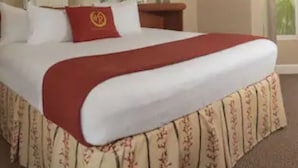 Un lit bien fait, recouvert d’une couverture rouge et d’une housse à motifs floraux, une table de chevet en bois et une fenêtre donnant sur des palmiers.