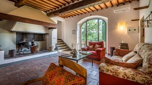 A cozy living room with a fireplace, a red sofa, a patterned armchair, and a large window offering a view of greenery.