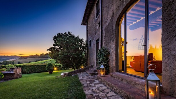 A stone building with large windows overlooking a lush garden and rolling hills.