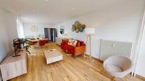 A modern living room with a wooden floor, a red sofa, a white armchair, and a dining area with a table and chairs. - Bail Mobilité - Finkwiller (Strasbourg)
