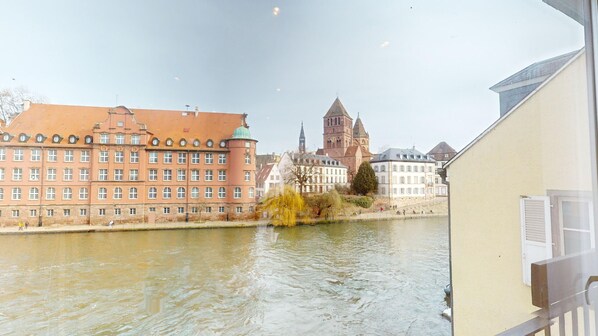 A river with buildings on the opposite bank, including a prominent tower and a red building with multiple windows.