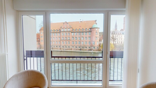A room with a large window overlooking a river and a historic building. - Bail Mobilité - Finkwiller (Strasbourg)