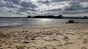 A beach with footprints in the sand, a body of water, and a distant shoreline with buildings. - Quick walk to private beach from this brand new 3-bedroom cottage (Wareham)