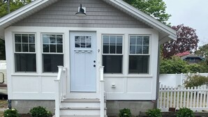 A small, single-story house with a white exterior, a front door, and a small porch with steps. - Quick walk to private beach from this brand new 3-bedroom cottage (Wareham)