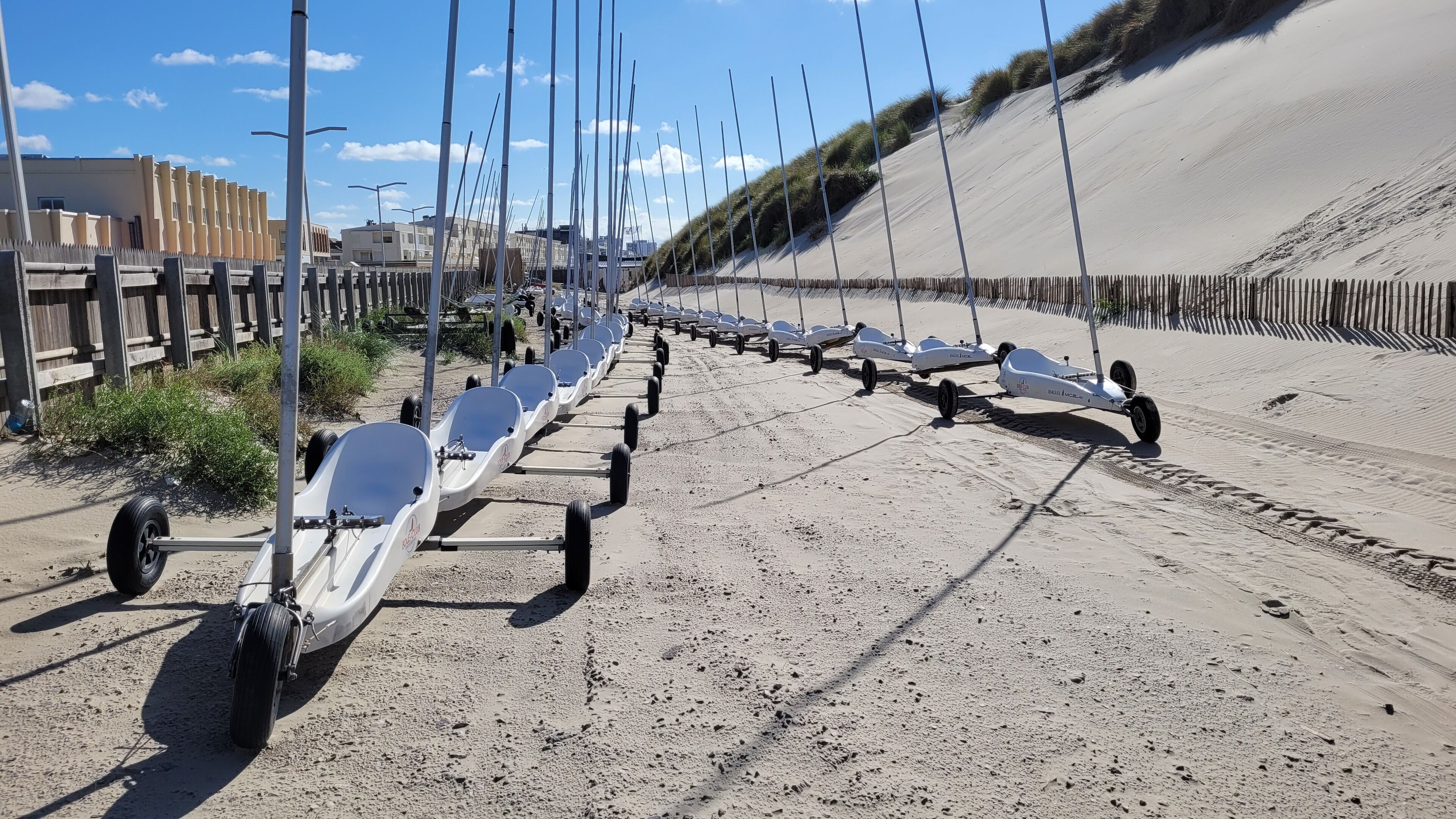 A row of stationary watercraft on a sandy beach with a wooden fence and buildings in the background.