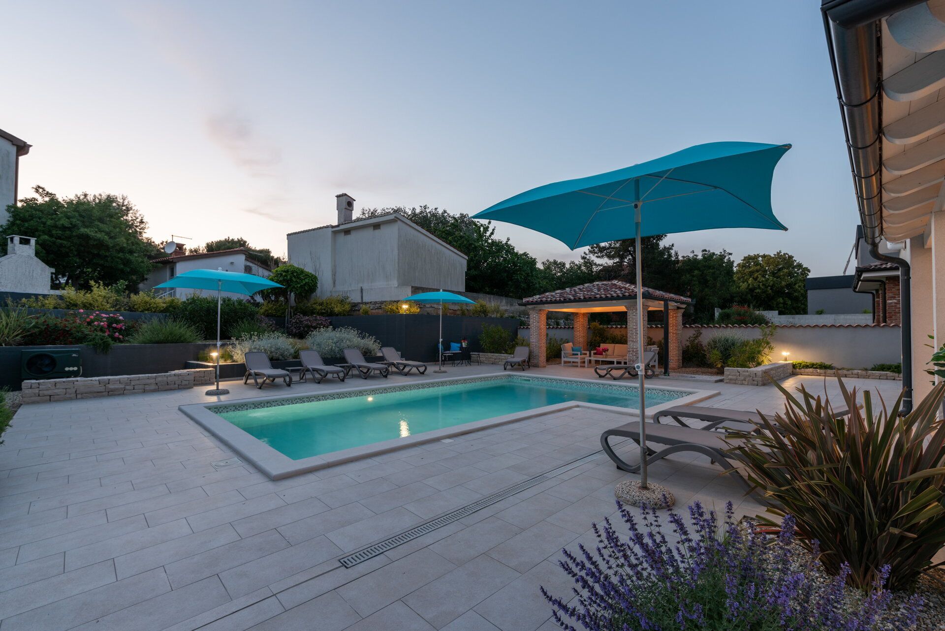 Une piscine entourée de chaises longues et de parasols, elle-même bordée d’un jardin aménagé et d’un bâtiment en briques.