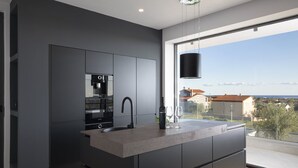 A modern kitchen with a built-in island, a coffee machine, and a view of the city through large windows. - Luxury Villa Maroon with Pool (Medulin)