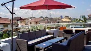 Eine Dachterrasse mit Korbmöbeln, einem roten Sonnenschirm und Blick auf die umliegenden Gebäude.