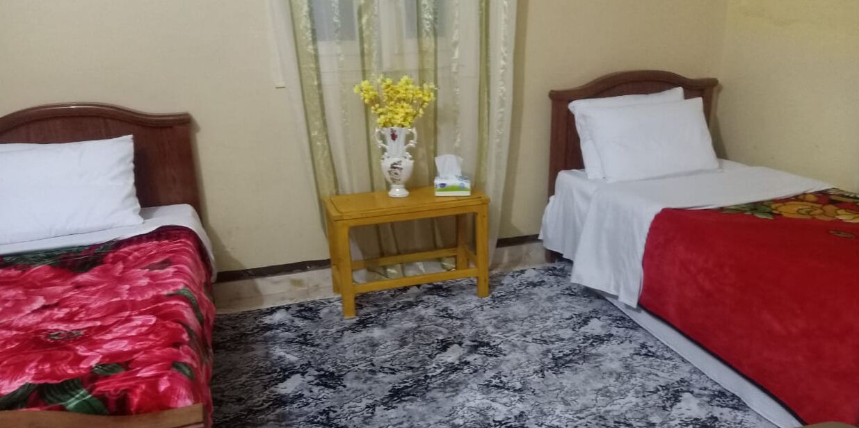 A hotel room with two single beds, a wooden nightstand with a vase of flowers, and a patterned carpet.