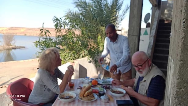 A group of people dining outdoors with a table set with food and drinks, a body of water, and greenery in the background. - tut amun guest house (Abu Simbel)