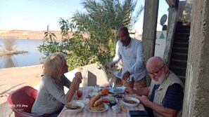 Un grupo de personas cenando al aire libre con una mesa dispuesta con comida y bebidas, un cuerpo de agua y follaje al fondo.