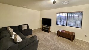 A living room with a dark-colored sofa, a television on a stand, a wooden bench, and a window with a view of a fenced area. - 2 Bdr 1 Bath Mountain View House (Tucson)