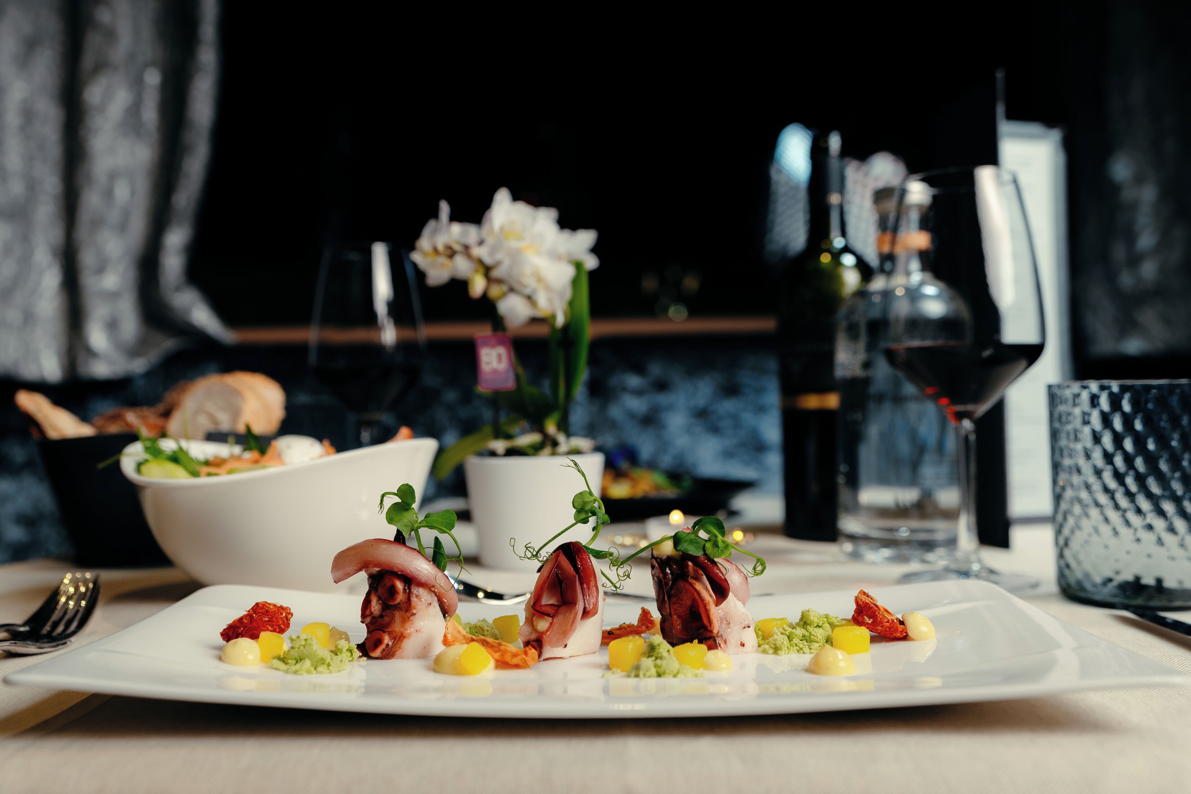 A dining table with a white plate of food, a bowl of salad, and a bottle of wine.