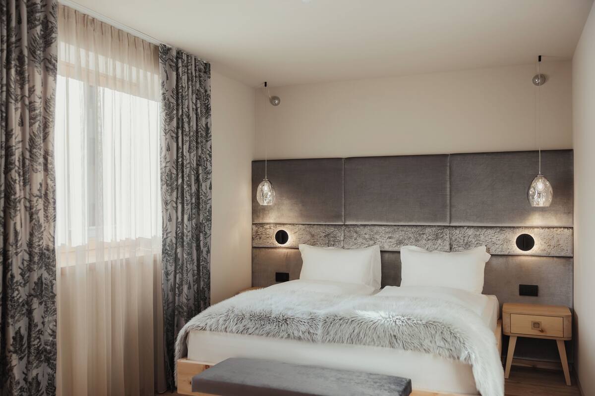 A modern bedroom with a large bed, two bedside lamps, a nightstand, and a window with sheer curtains.