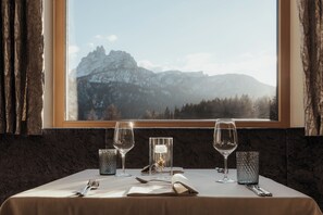 A table set for two with wine glasses, a candle, and a menu, overlooking a mountainous landscape. - Hotel Tofana Cortina  (Cortina d'Ampezzo)