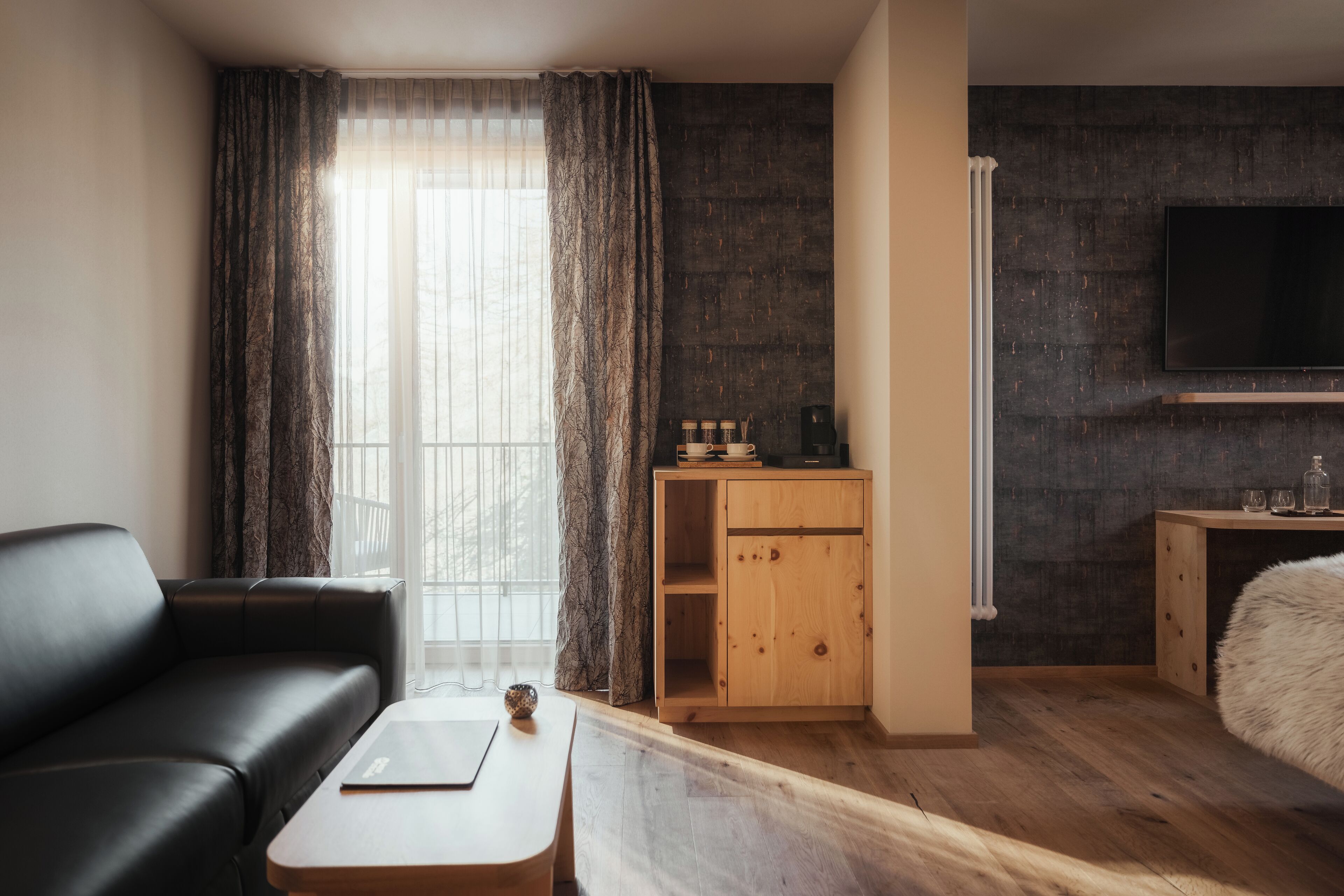 Una habitación de hotel moderna con un sofá de cuero negro, una mesa de centro de madera, un armario de madera y un televisor de pantalla plana grande montado en la pared.