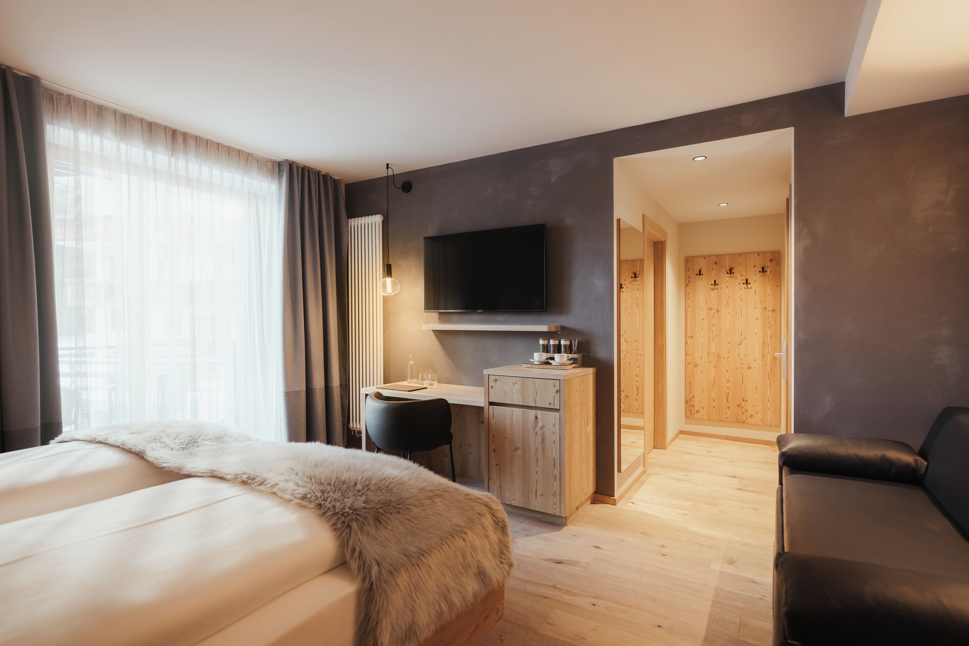 A modern hotel room with a large bed, a desk with a chair, a television, and a wooden wardrobe.