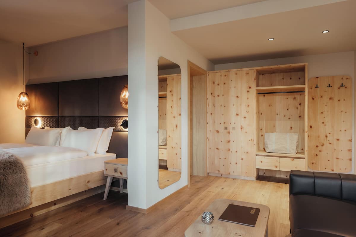 A modern hotel room with a large bed, a desk, a mirror, a seating area, and wooden storage units.
