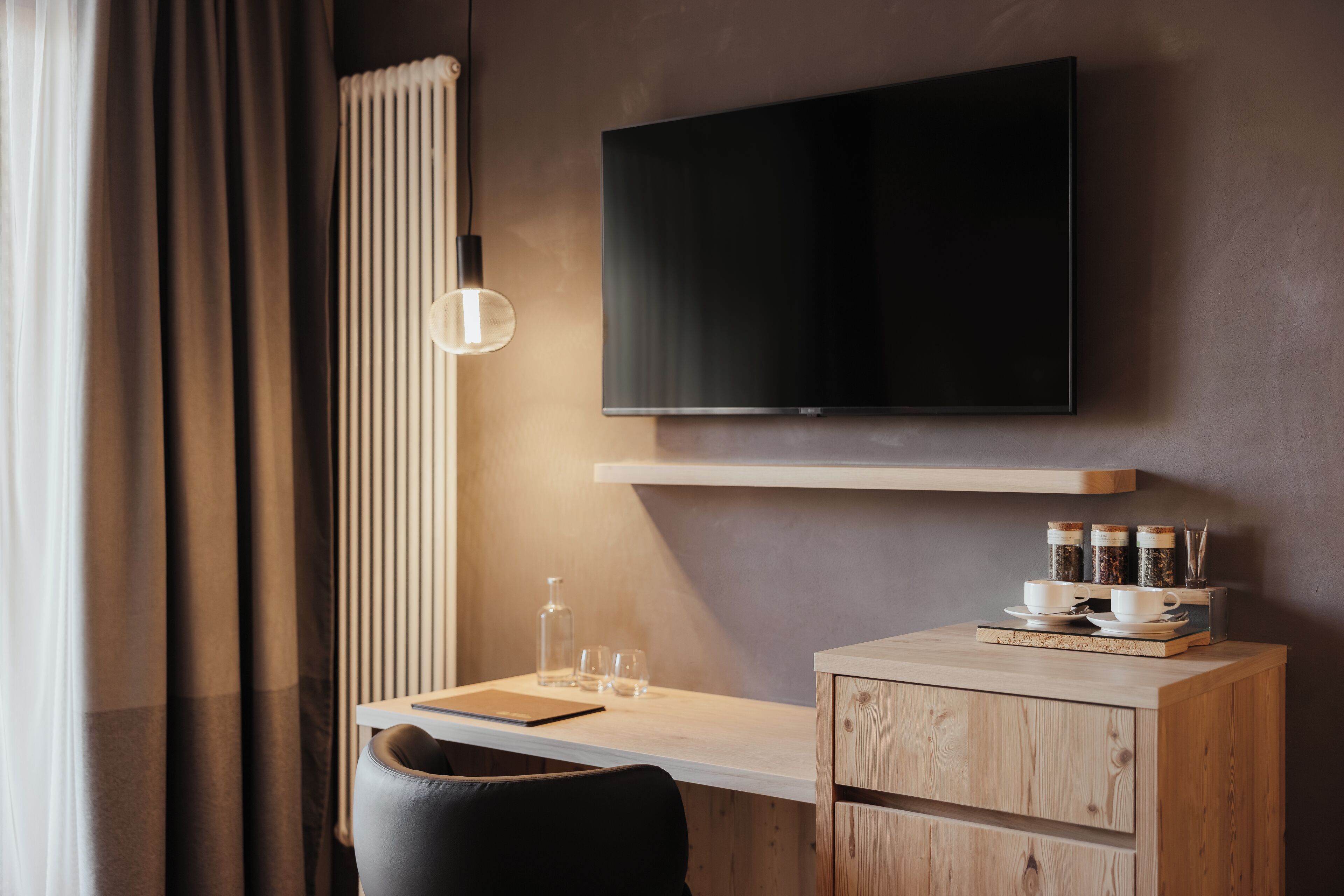 Una habitación de hotel moderna con un televisor de pantalla plana montado en la pared, un escritorio de madera con una computadora portátil, una silla y un mueble bajo.