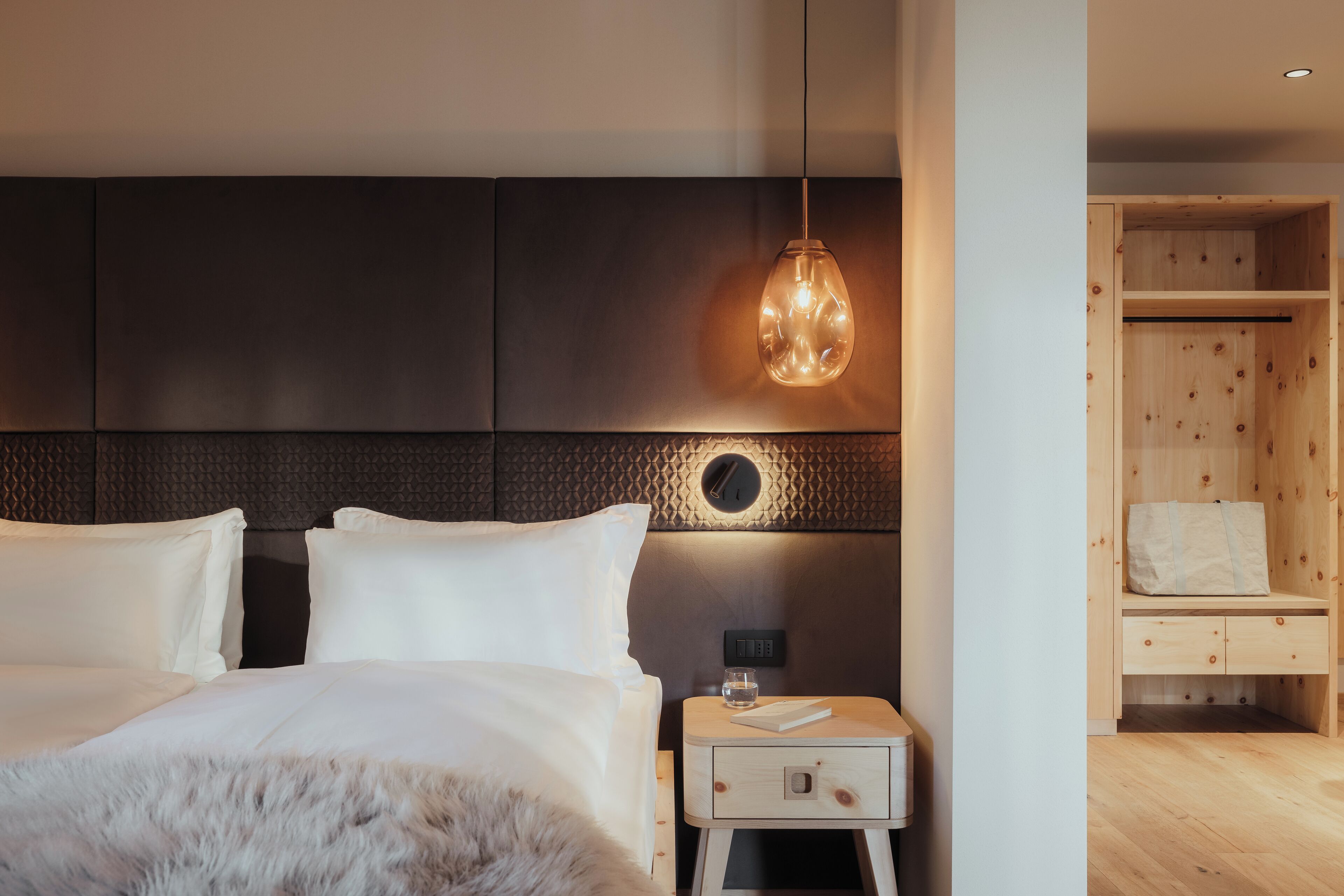 A modern hotel room with a large bed, a nightstand, a wooden wardrobe, and a pendant light.